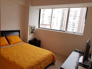 Een slaapkamer met een bed, een nachtkastje, een televisie en een raam met uitzicht op de stad.