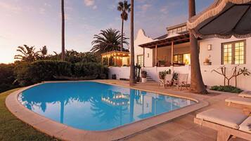 Relax by the serene pool surrounded by lush greenery and palm trees, with a patio area offering comfortable outdoor seating.