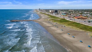 A coastal beach with a long stretch of sand, a pier extending into the sea, and a road running parallel to the beach.