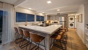 A modern kitchen with a central island, bar stools, a built-in oven, and a washing machine.
