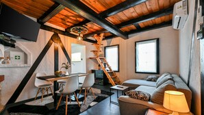 Una sala de estar acogedora con techo de madera, un comedor con mesa y sillas, un sofá con lámpara, y una ventana que deja entrar luz natural.