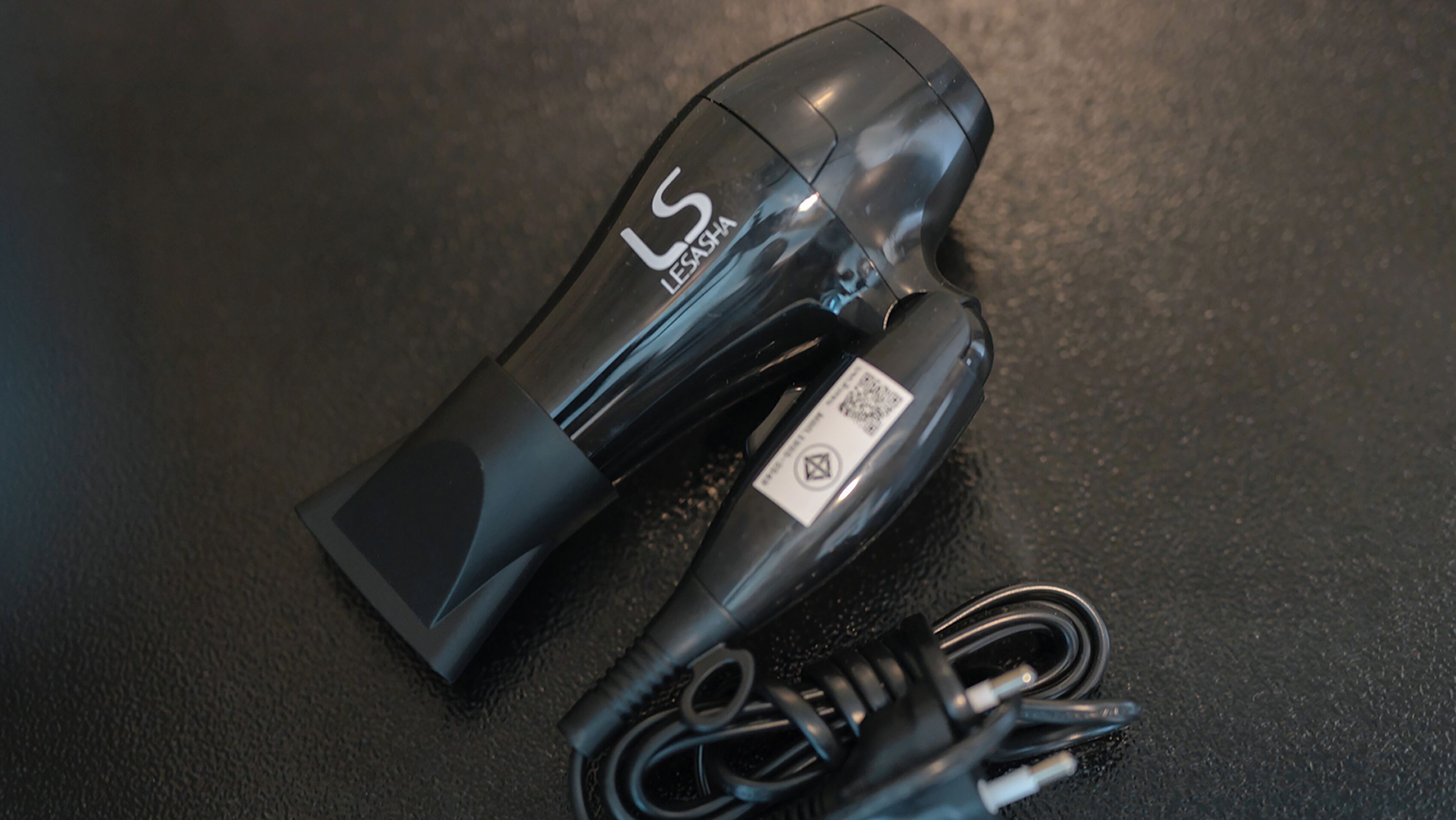 A black hair dryer with a cord and a power adapter on a textured surface.