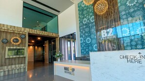 A modern hotel lobby with a reception desk labeled "B2 Premier" and a "Check-In Here" sign. There are clocks on the wall and a "Wake Up" sign on the reception desk.