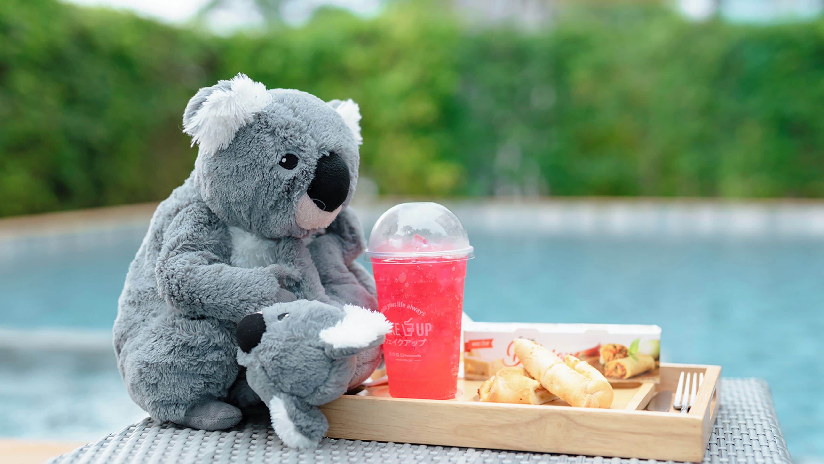 A plush koala toy sitting next to a tray with a drink and food items.