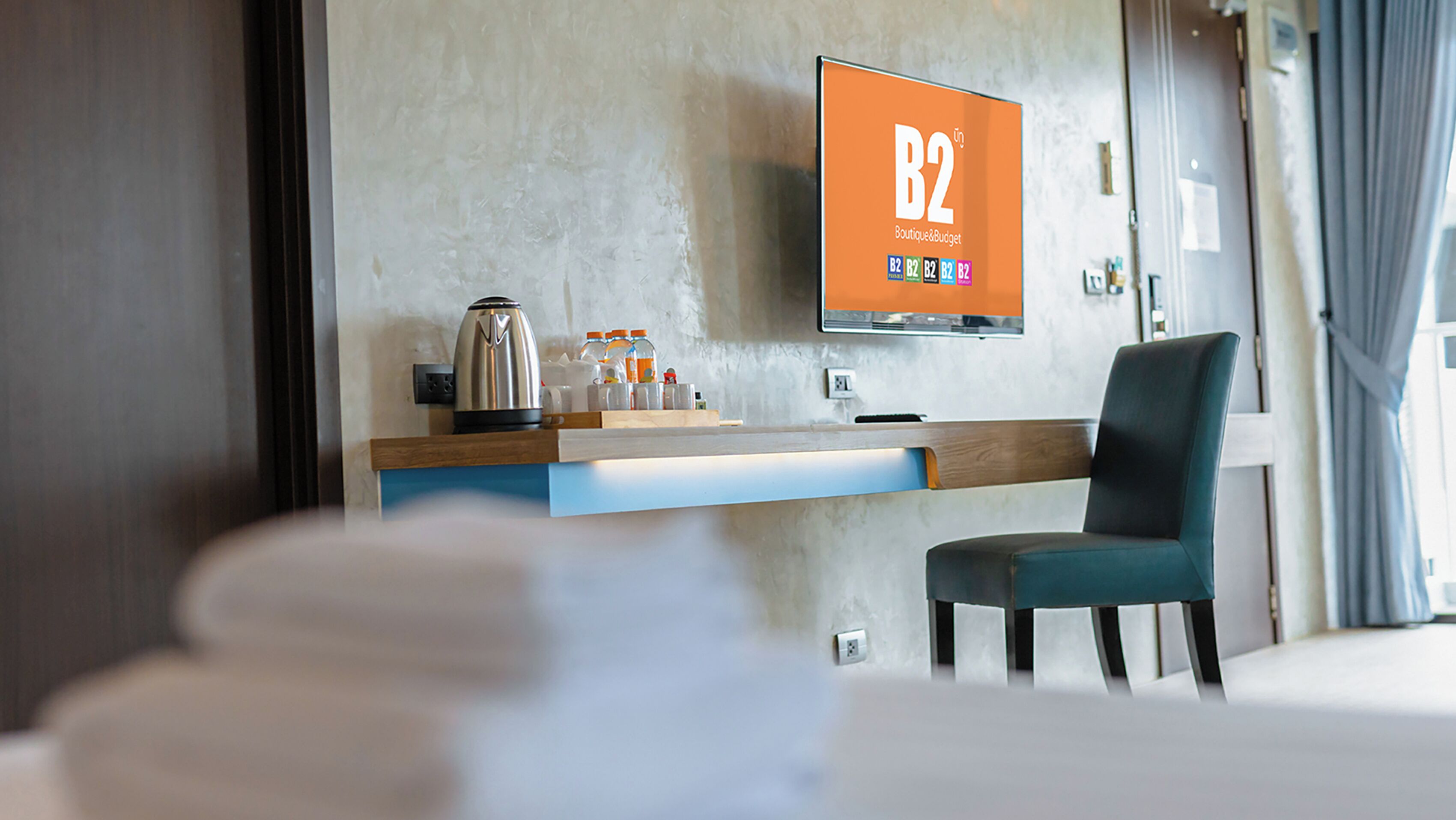 Et moderne hotelværelse med et fladskærms-TV, der viser 'B2 Boutique & Budget' på væggen, et træsidebord med elkedel og flasker, en blå stol og en hvid seng.