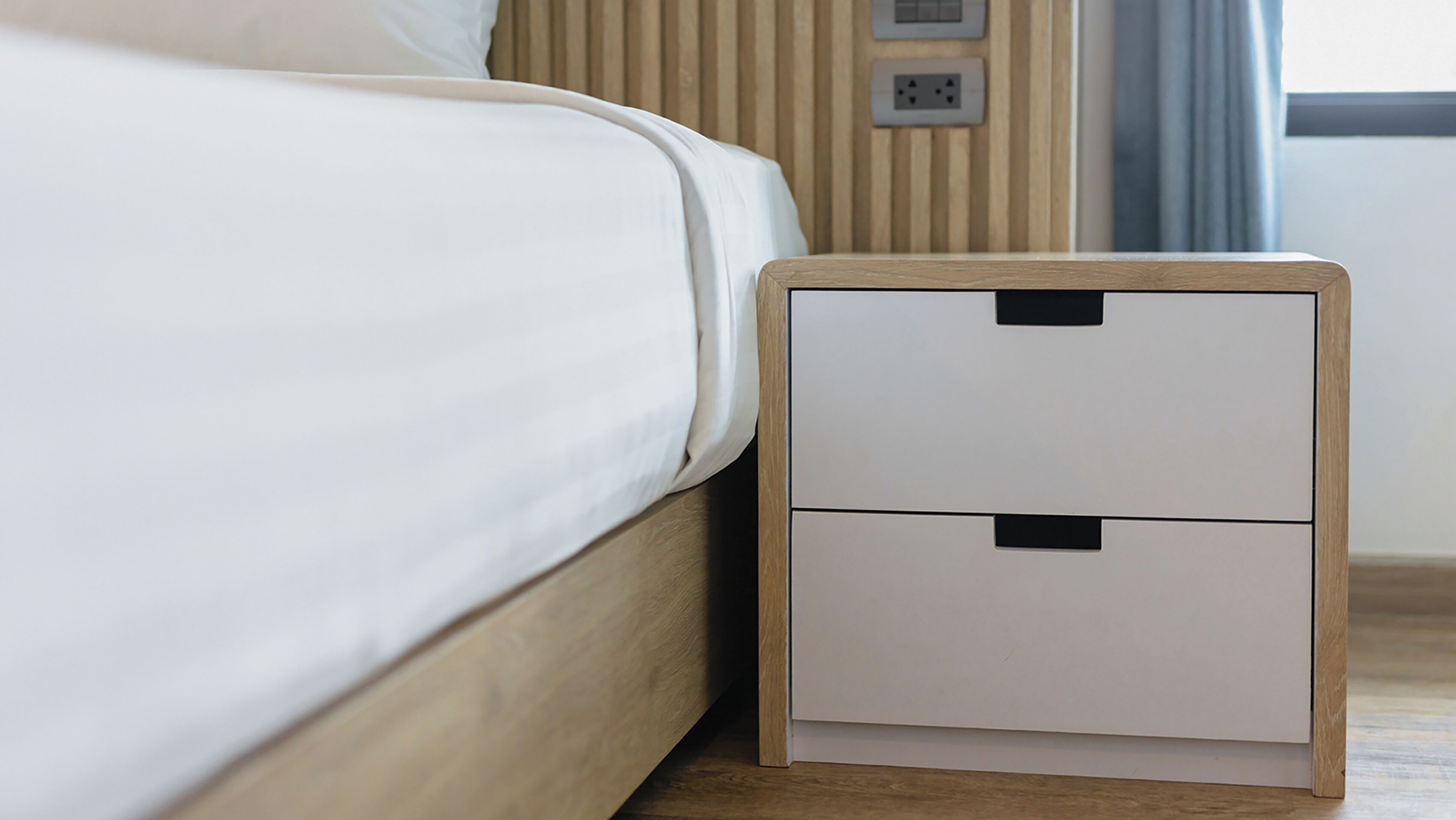 A neatly made bed with a wooden headboard and a white mattress. A small wooden nightstand with two drawers is placed beside the bed.