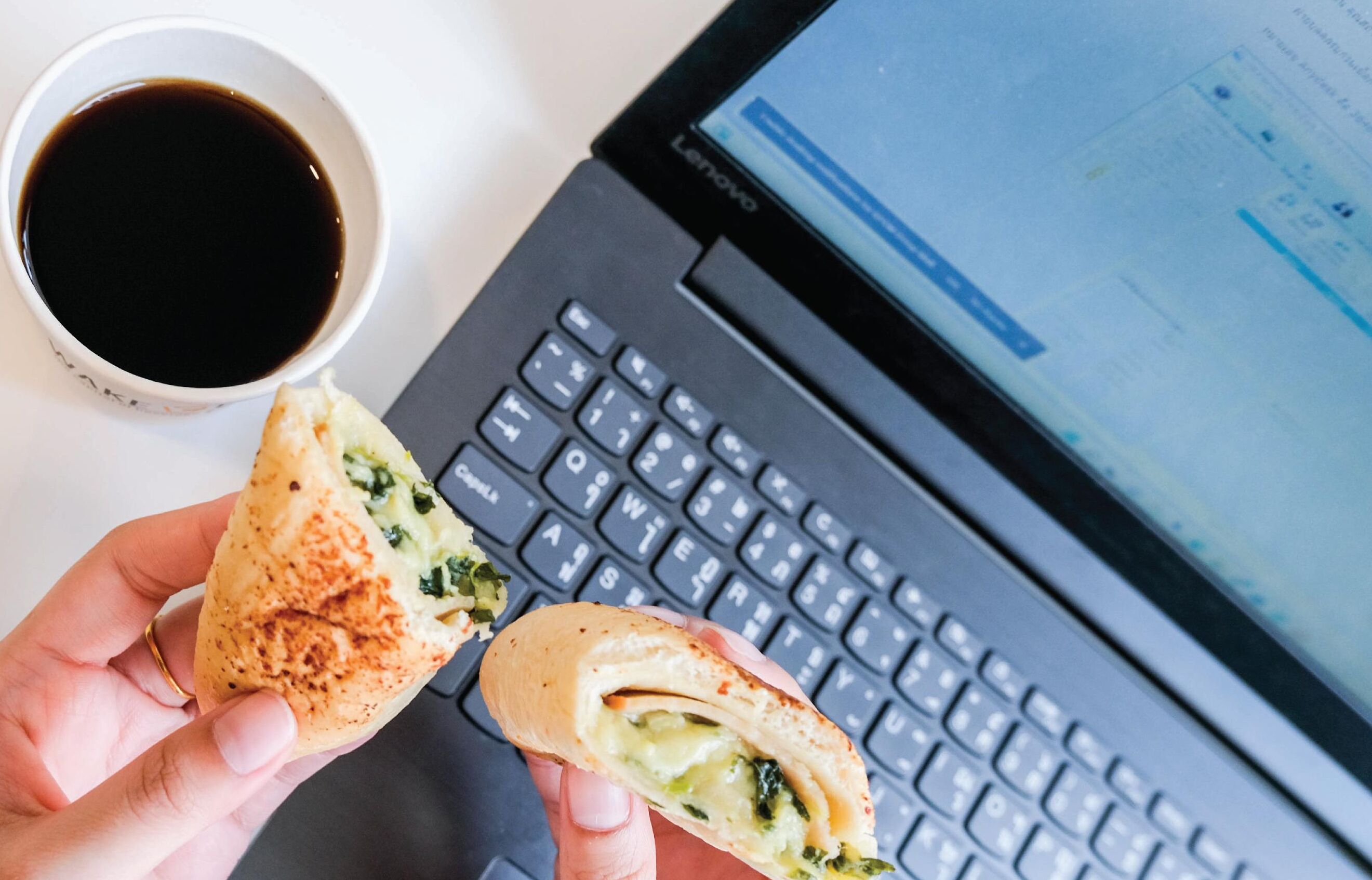 Una computadora portátil con teclado y pantalla mostrando texto, y una mano sosteniendo un sándwich con vegetales verdes y queso visible.