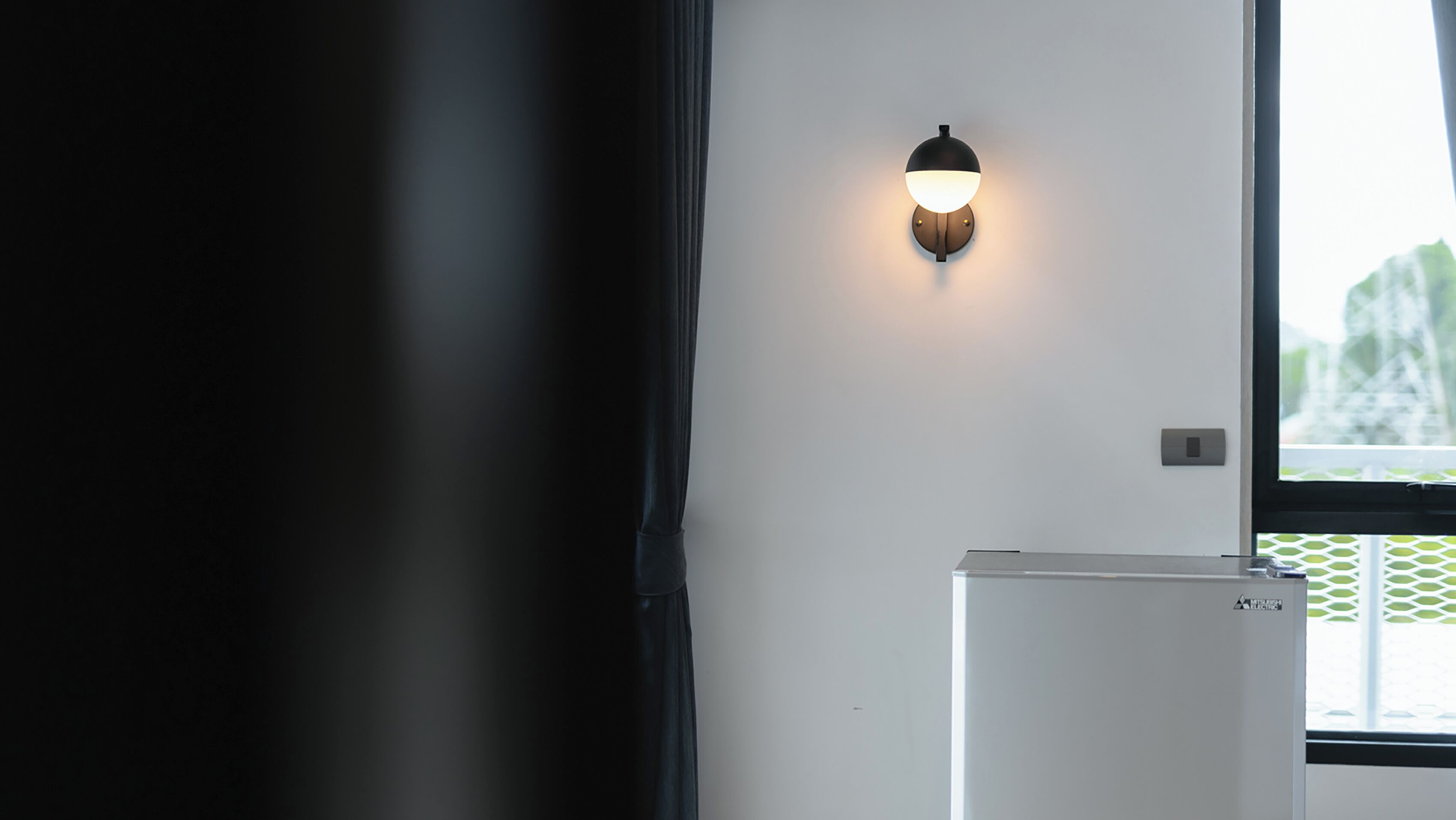 Una habitación con pared blanca, una lámpara fijada a la pared, una ventana con vista a follaje y un electrodoméstico blanco.
