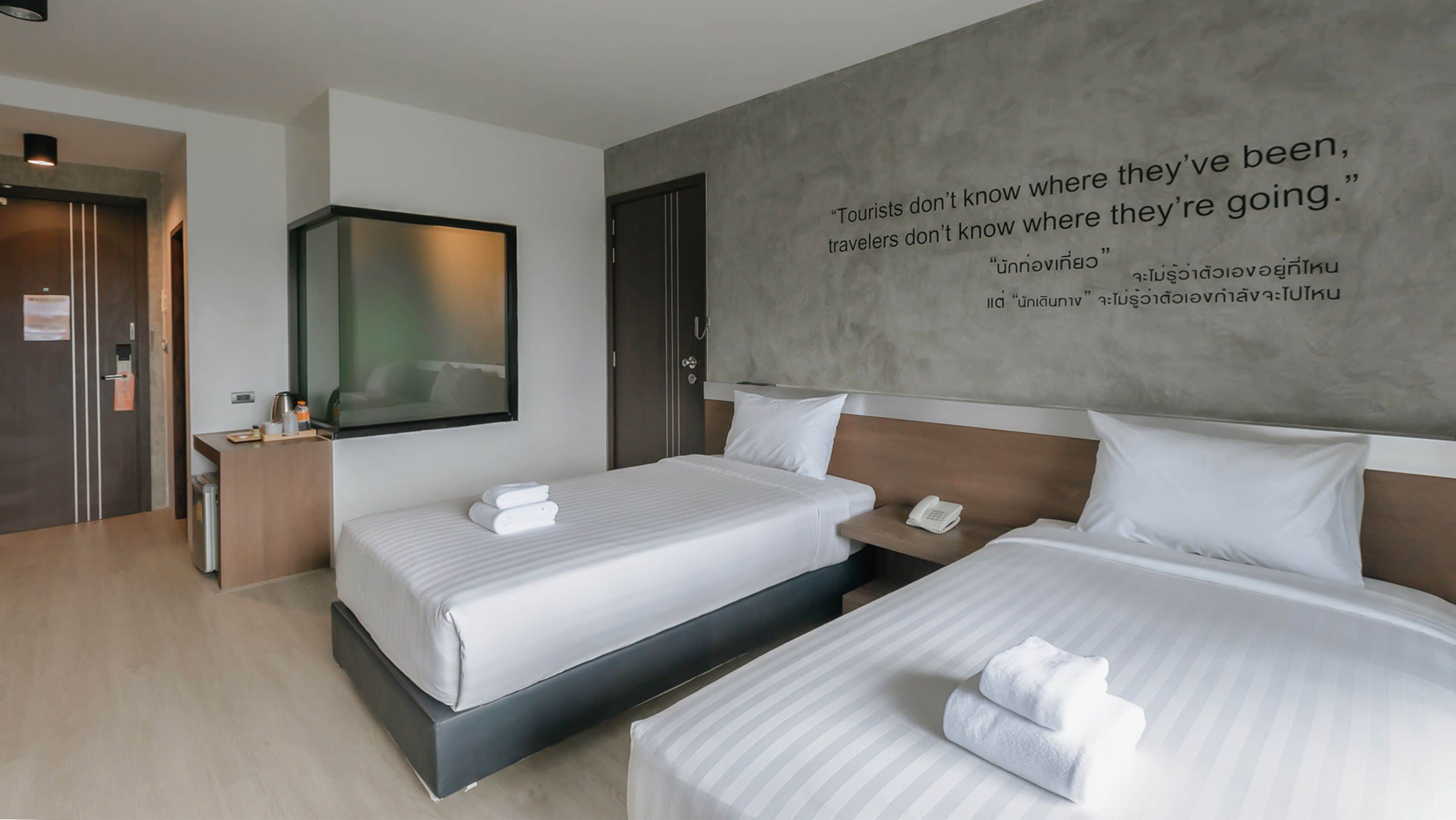 A modern hotel room with two beds, a bedside table with a phone, and a wall with a motivational quote.