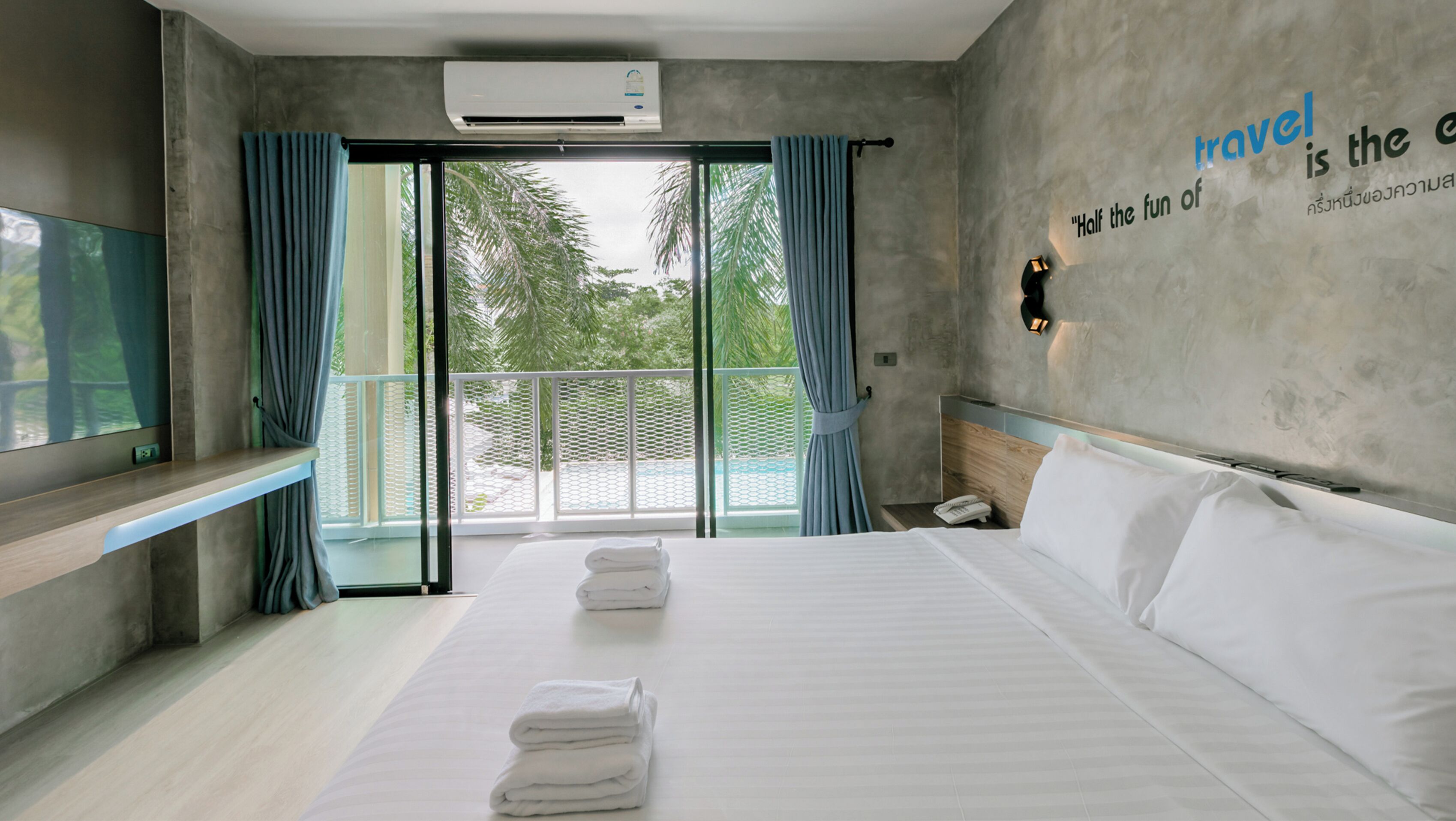 A modern hotel room with a bed, a TV, a balcony with curtains, and a wall with a travel quote.