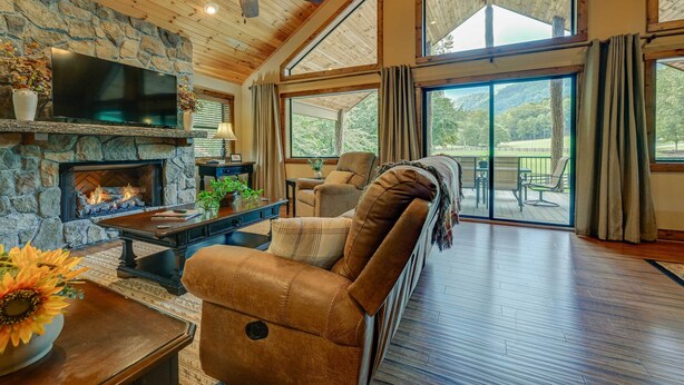 A cozy living room with a stone fireplace, leather sofa, and large windows offering a view of the outdoors. - 28 Acres: Home w/ Barn & Fire Pit Near Franklin! (Otto)