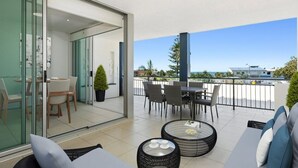 A modern balcony with a glass door leading to a dining area, furnished with a table and chairs, and a seating area with a sofa and coffee table. - Ocean View Haven at Paradiso (Kingscliff)