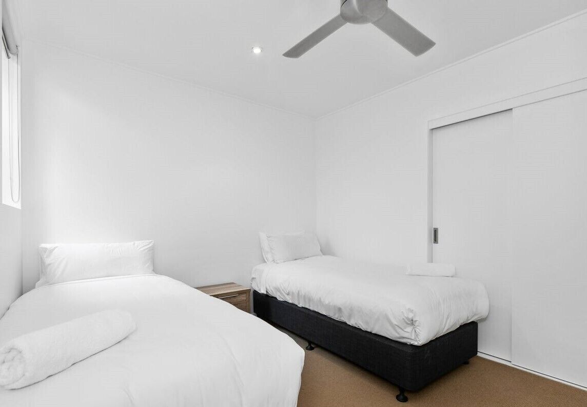 A room with two beds, a ceiling fan, a wardrobe, and a small bedside table.