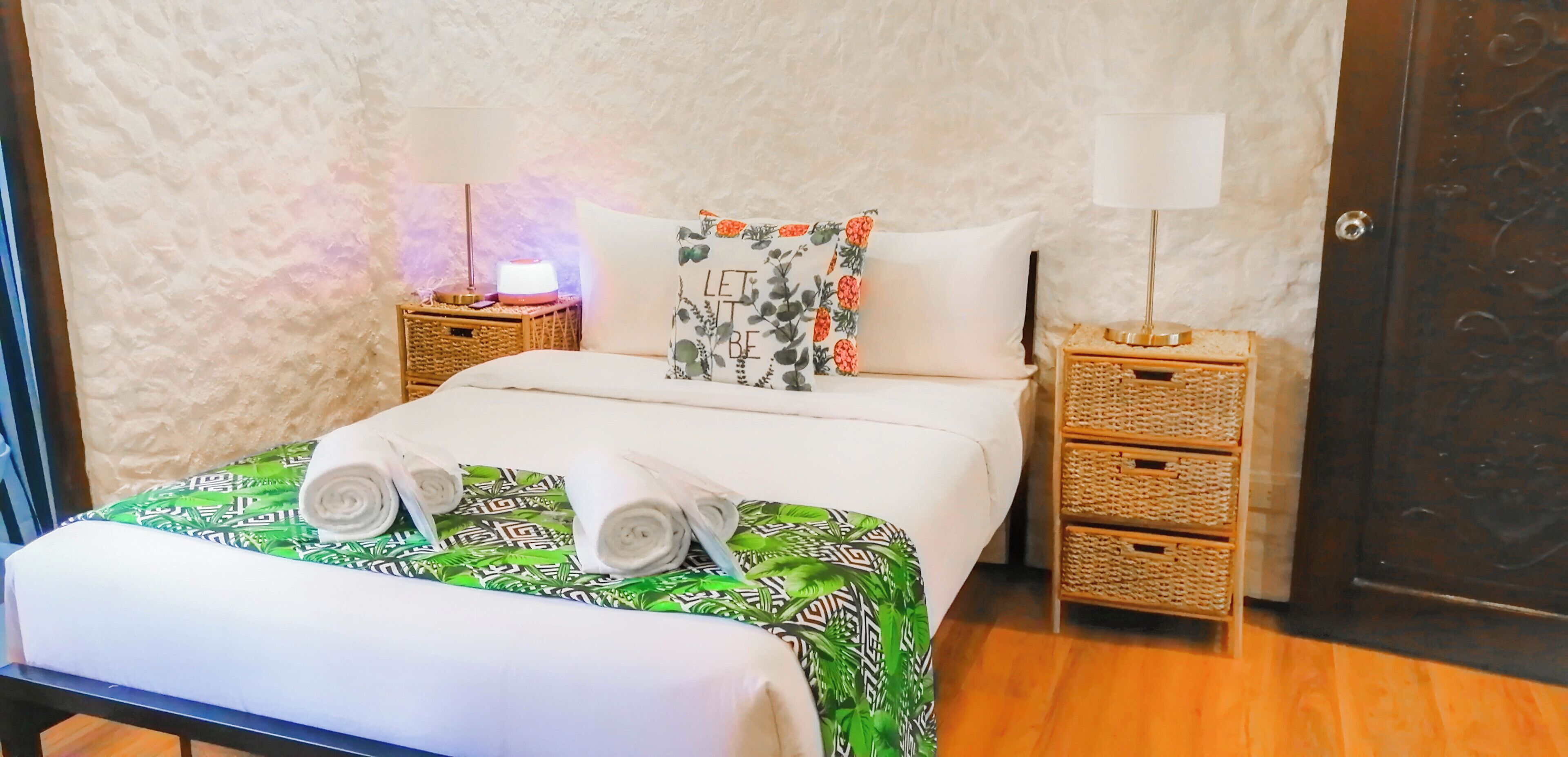 A neatly made bed with a green patterned bedspread, two rolled towels, a floral pillow, and a bedside lamp on a wooden nightstand.
