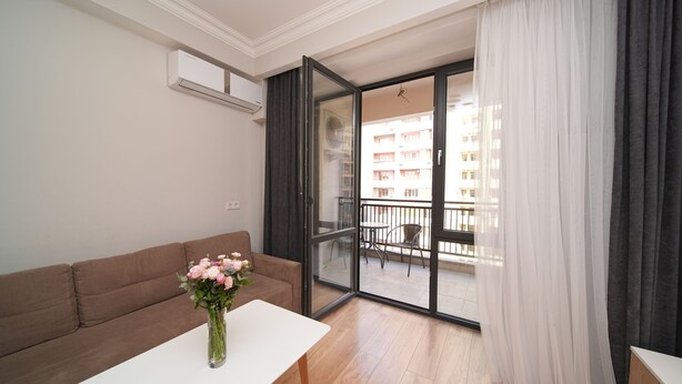 A modern apartment with a sofa, a small table with a vase of flowers, a sliding glass door to a balcony, and a white air conditioning unit on the wall.