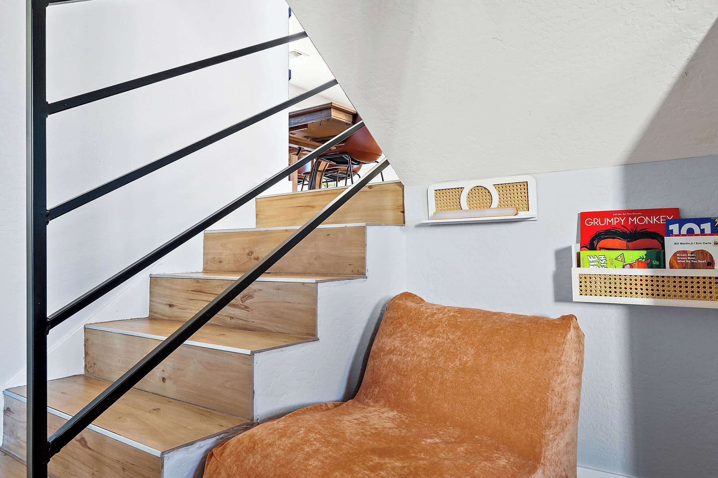Un escalier avec des marches en bois et des rampes noires, un fauteuil rembourré et une étagère fixée au mur, ornée de livres et d’un objet décoratif.