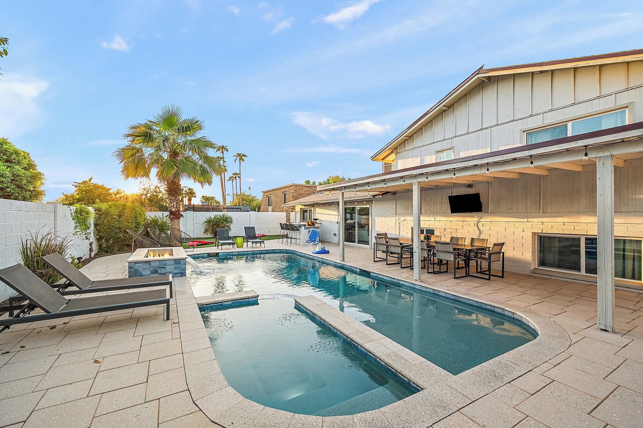 Piscina con sillas de descanso, una fogata y un patio techado con mobiliario exterior.