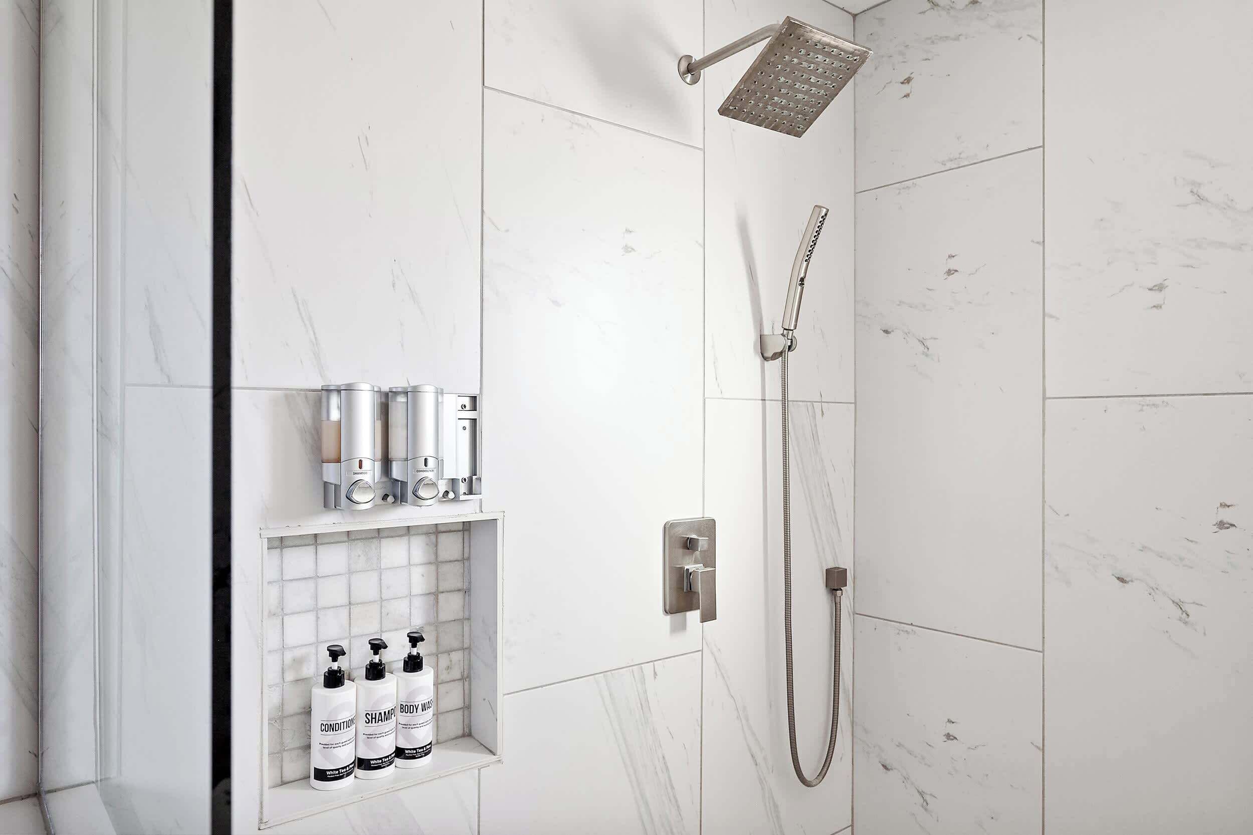Un área de ducha moderna con un cabezal de ducha manual y otro empotrado en la pared. Hay tres botellas de champú y acondicionador en un estante debajo de la ducha.