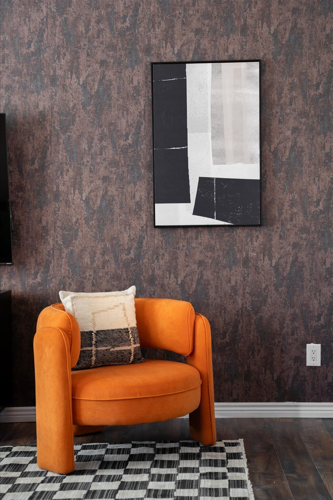 Una stanza con un muro marrone con texture, un quadro astratto in bianco e nero, un divano arancione con un cuscino decorato e un tappeto a scacchi bianchi e neri.