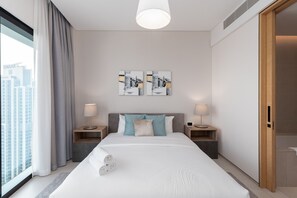 Une chambre d’hôtel moderne avec un grand lit, deux tables de chevet avec des lampes, une vue sur la ville et des œuvres d’art au mur.
