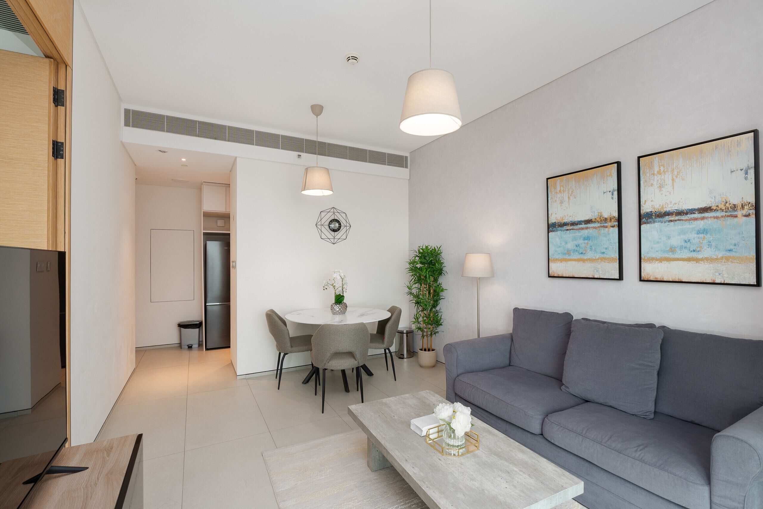 A modern living room with a grey sofa, a round dining table with chairs, a coffee table with a vase of flowers, and two wall-mounted lamps.