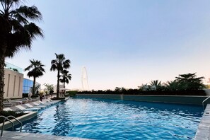 Una piscina con sillones de descanso y palmeras, una noria al fondo y edificios modernos.