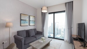 Una sala de estar moderna con un sofá gris, una mesa de centro, una lámpara y un televisor. Hay dos cuadros en la pared y un ventanal con cortinas.