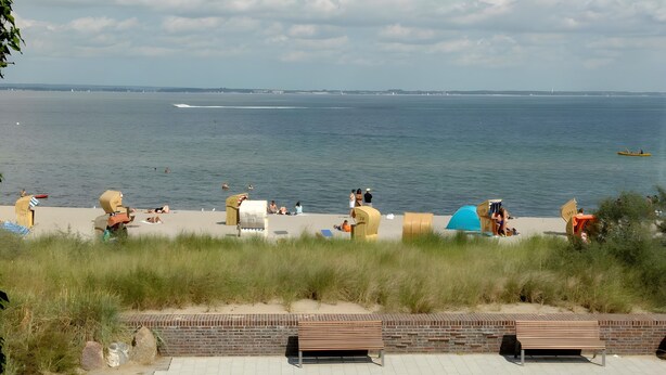 Una playa con gente relajándose, un banco y vista al mar.