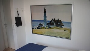 A bedroom with a bed, a painting of a lighthouse, and a black bag on the wall.