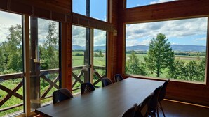 A wooden dining area with a table and chairs, large windows offering a view of a green landscape and mountains. - Biei san-ai-no-oka Villa Shion (Biei)