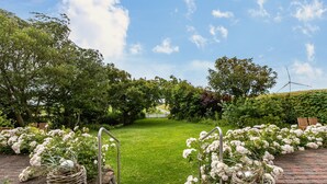 A paved pathway leading to a grassy area with a metal gate, surrounded by lush greenery and white flowers. - Apartment 'Fewo Samson' with Private Terrace, Shared Garden and Wi-Fi (Emmelsbüll-Horsbüll)