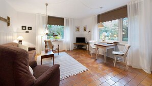 Ein Wohnzimmer mit einer Couch, einem Esstisch mit Stühlen, einem Fernseher und einem Fenster mit Vorhängen.