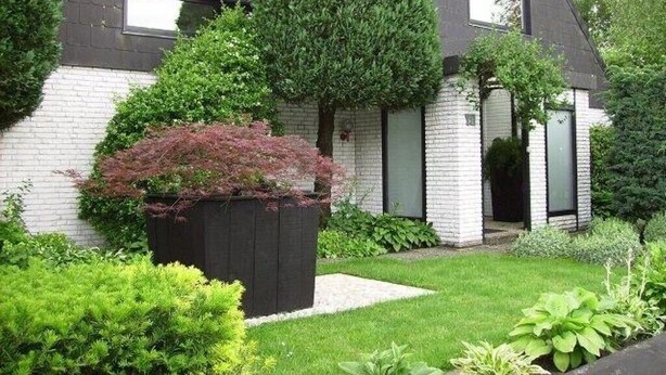 Ein modernes Haus mit einem geneigten Dach, einer Ziegeläußeren und einem gepflegten Garten mit verschiedenen Sträuchern und einem großen Baum.
