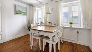 A dining area with a wooden table and white chairs, a white radiator, a window with curtains, and a painting on the wall. - Apartment 'Fewo Luna' with Private Terrace, Shared Garden and Wi-Fi (Emmelsbüll-Horsbüll)