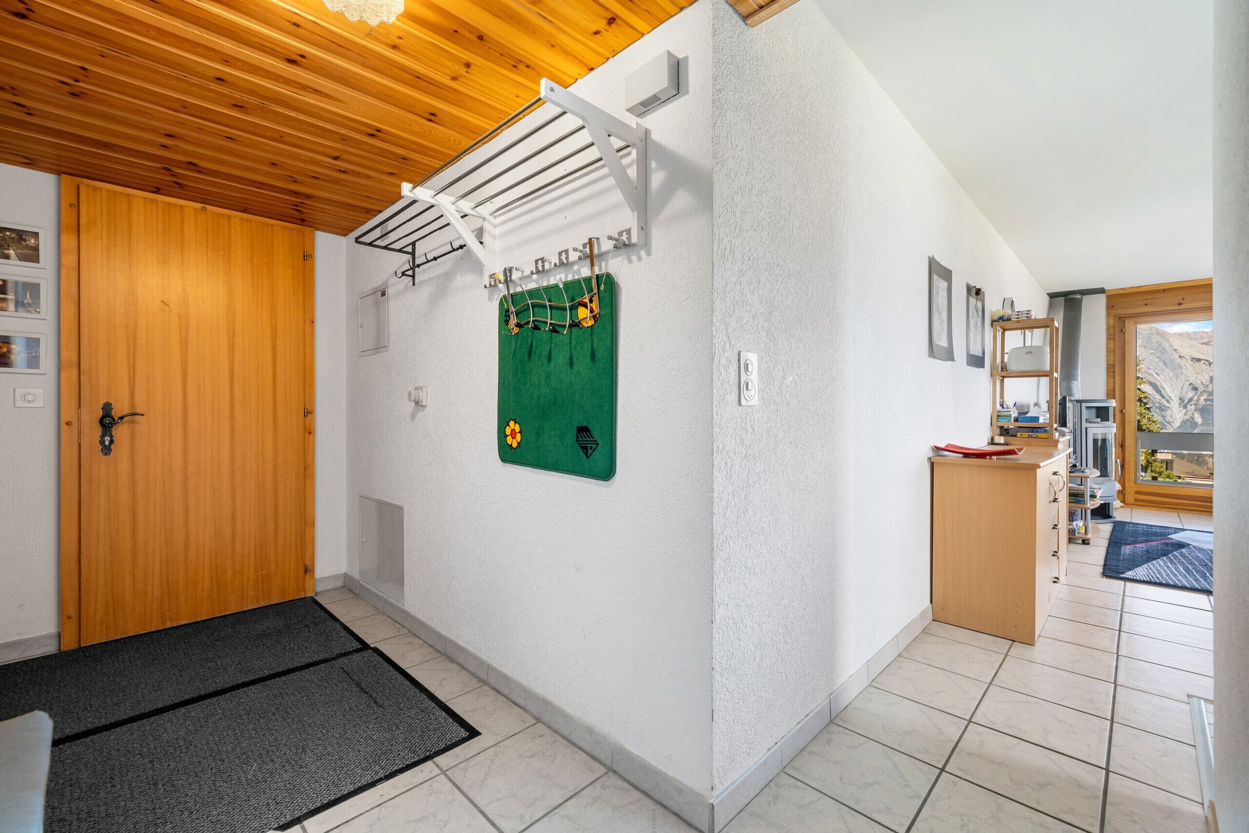 A hallway with a wooden door, a green wall-mounted board, a white shelf, and a view of a mountain through a window.