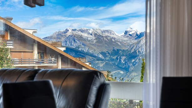 A modern interior with a leather sofa and a view of snow-capped mountains through a window. - Les Hauts-de-Nendaz A A0 - 5 pers - ski In&Out (Haute-Nendaz)