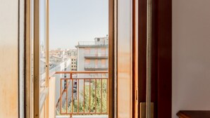 Blick aus einem Fenster mit Metallbalkongeländer, blauem Himmel und Gebäuden im Hintergrund.