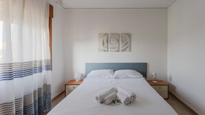 Ein ordentlich bezogenes Bett mit weißen Leinentüchern, einem blauen Kopfteil und einer Reihe Handtücher. Zwei Nachttische mit Lampen stehen bereit, und über dem Bett hängt ein Wandbild.