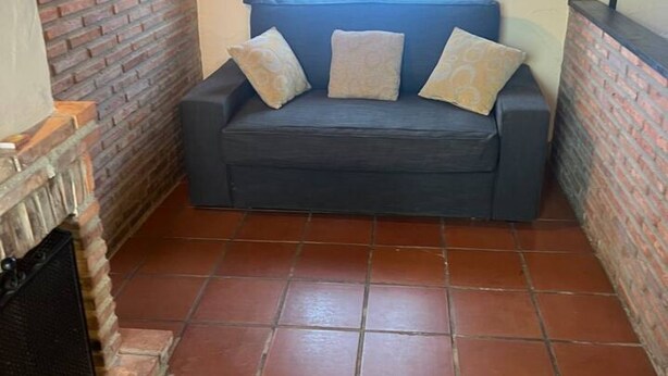 A corner room with a dark gray sofa, three patterned cushions, a tiled floor, and a brick wall. - 15A08 El Rosal (Llanes)