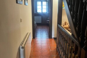 Un estrecho pasillo con un radiador, una ventana con cortinas y una escalera con barandilla.