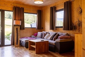 A cozy wooden room with a sofa, a small table, and a lamp. There are curtains on the windows, and a view of greenery outside.