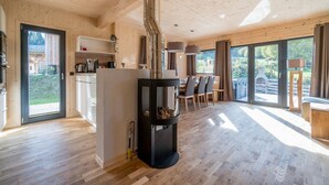 A modern wooden interior with a central wood stove, a kitchen area, and large windows offering a view of the outdoors. - Spacious chalet with sauna (Sankt Georgen am Kresichberg)