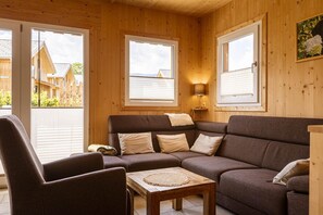 A cozy living room with a brown sofa, a wooden coffee table, and a lamp. There are large windows with blinds, and a view of a wooden fence outside.