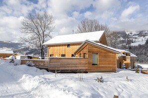 A wooden cabin with a sloped roof, surrounded by snow-covered ground and trees, set in a mountainous area.