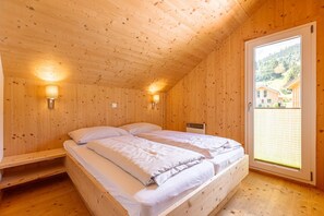 A wooden cabin bedroom with a bed, two wall-mounted lamps, and a large window with a view of a mountain and buildings.
