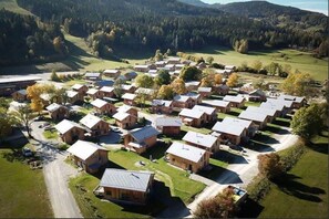 Eine Gruppe aus Holzgebäuden mit Satteldächern, angeordnet in einem Gittermuster, umgeben von grünen Feldern und Bäumen, mit einem Gebirgszug im Hintergrund.