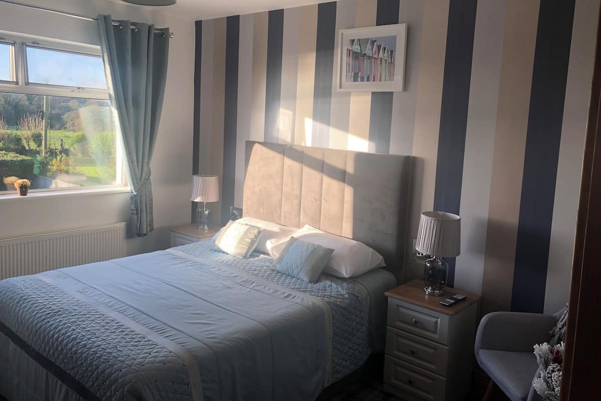 A bedroom with a large bed, a bedside table with a lamp, a chair, and a window with curtains.