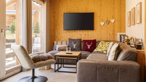 A cozy living room with a large flat-screen TV, a sofa, a chair, and a wooden wall with heart-shaped decorations.