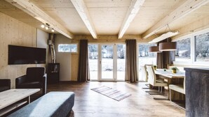 A spacious room with a wooden ceiling, a large window with curtains, a flat-screen TV, a dining area with a table and chairs, and a bench.