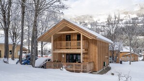 Een houten chalet met balkon, omringd door met sneeuw bedekte bomen en een bergachtige achtergrond.